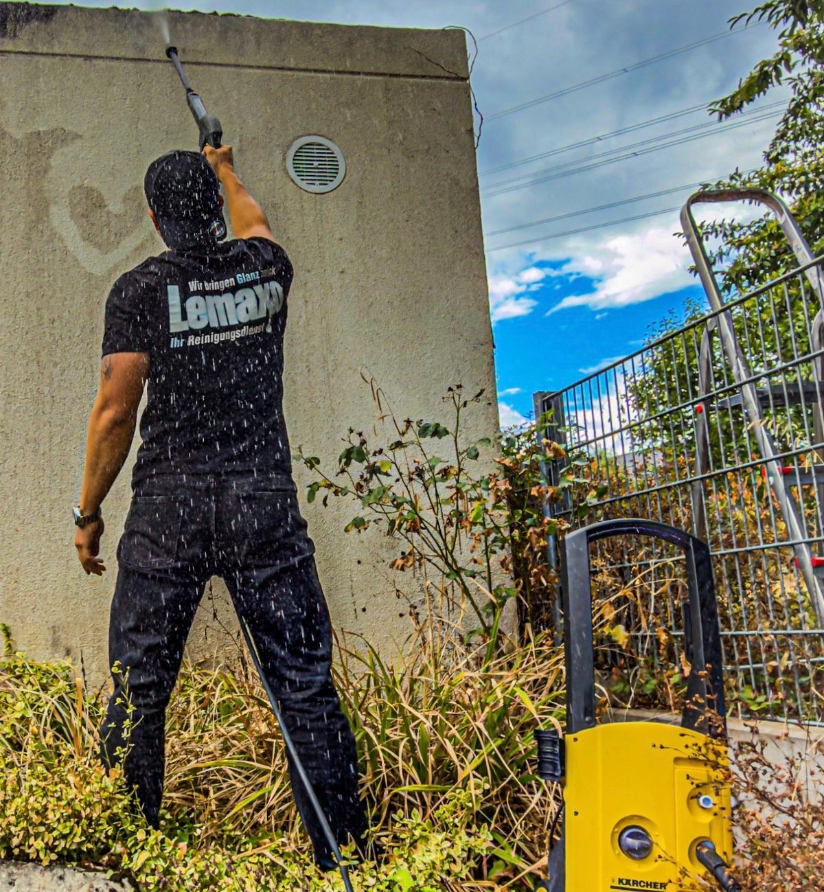 Man in black shirt using yellow pressure washer to clean concrete wall in sunny outdoor setting