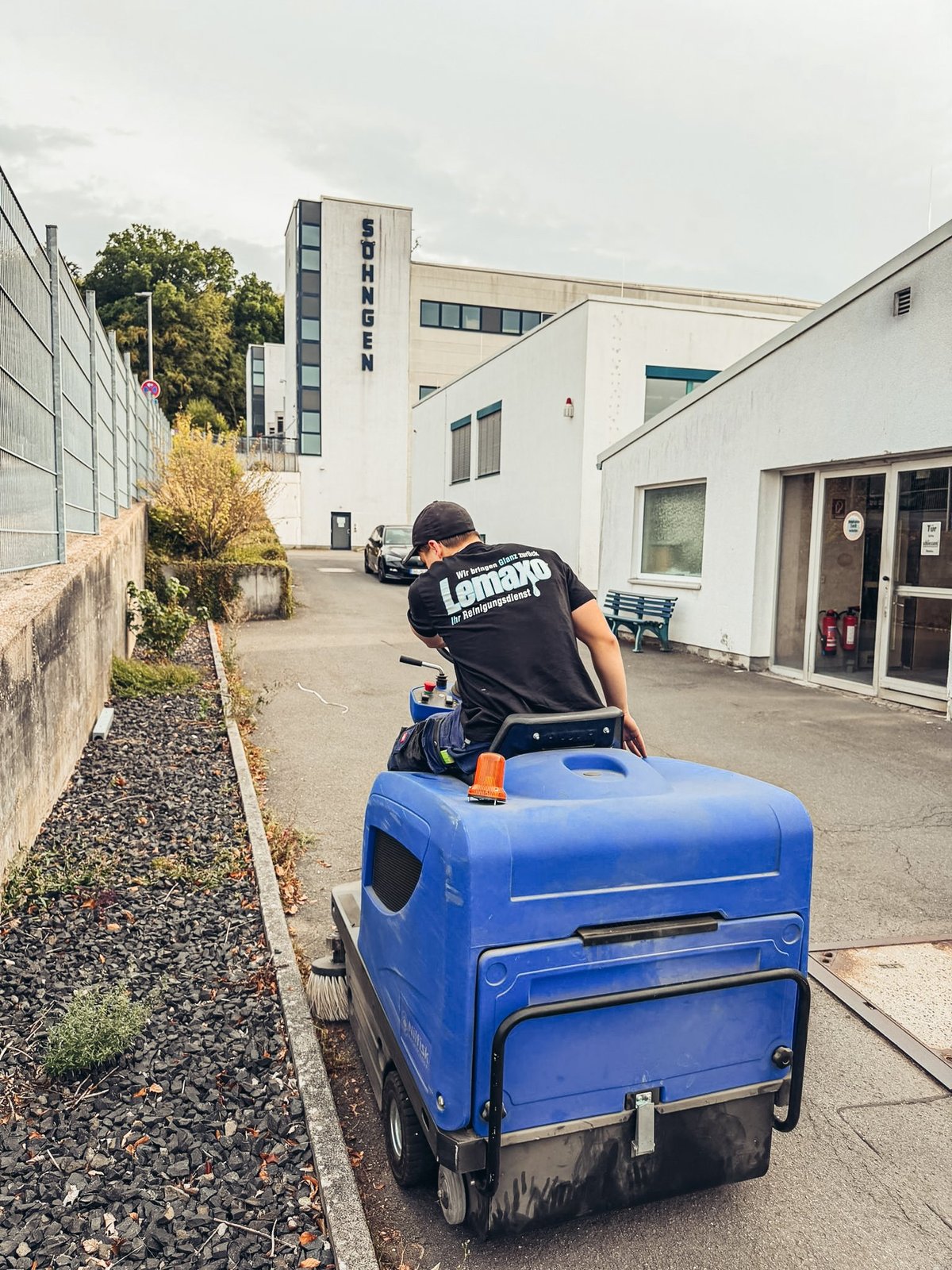 Worker riding a blue street cleaning machine in an industrial courtyard with modern buildings