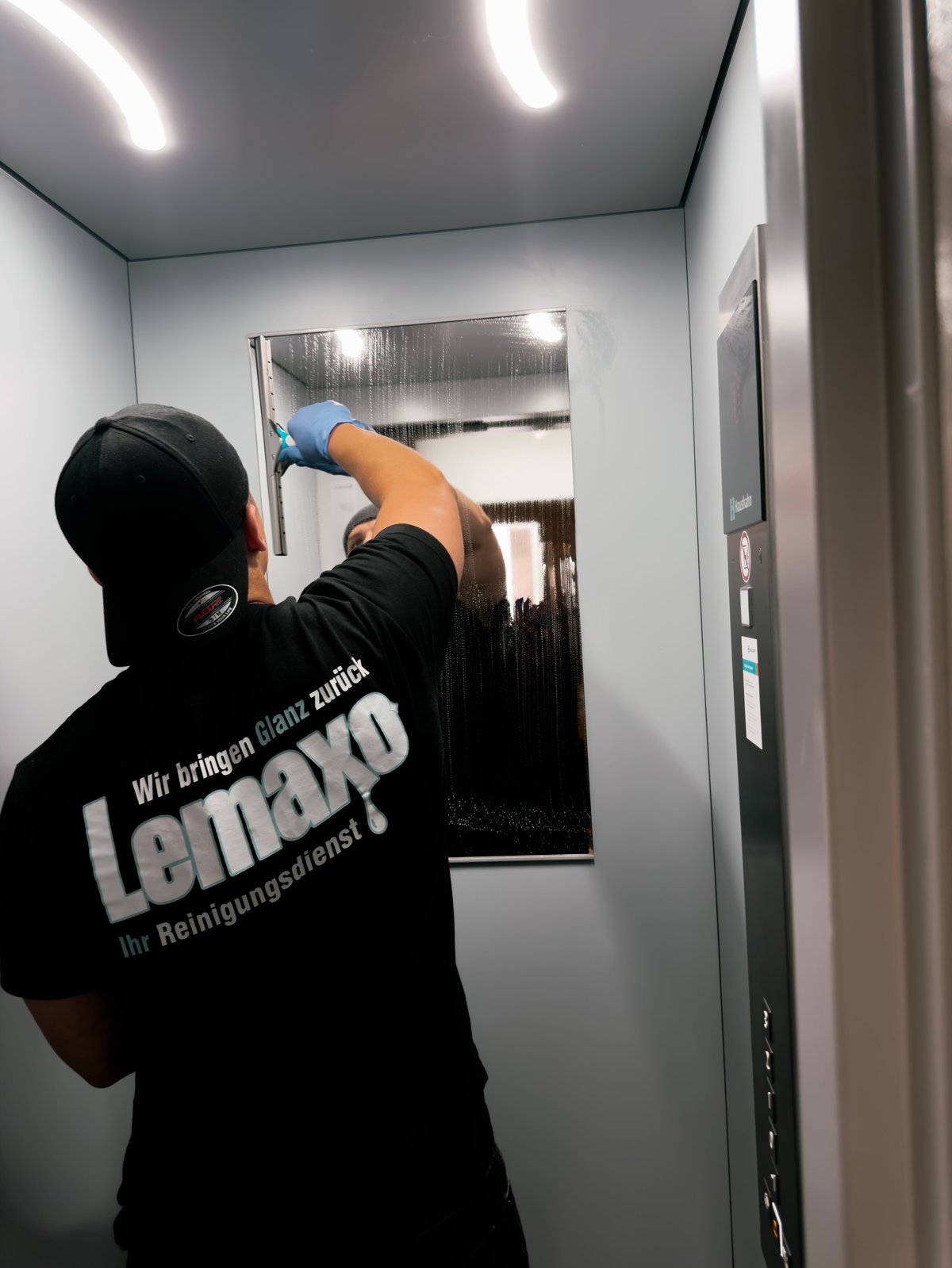Lemaxo-Mitarbeiter reinigt einen Spiegel, Schriftzug Wir bringen Glanz zurück auf dem Shirt